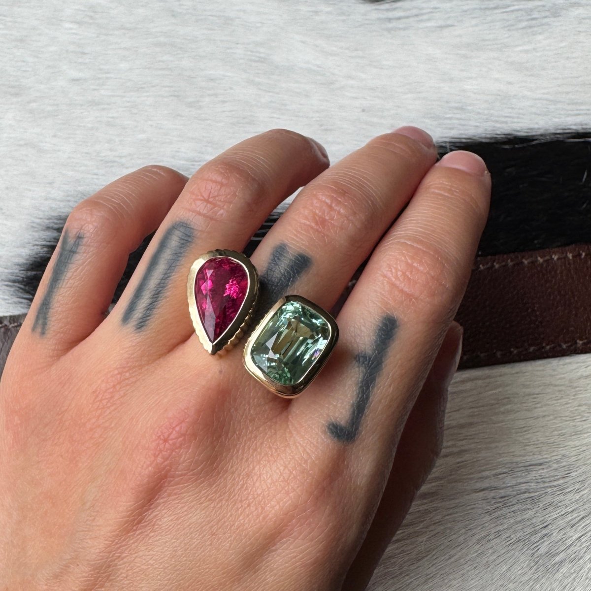 Hand wearing a gold ring with an elongated cushion-cut mint tourmaline and a pear-shaped pink red rubellite against a black and white cow print background