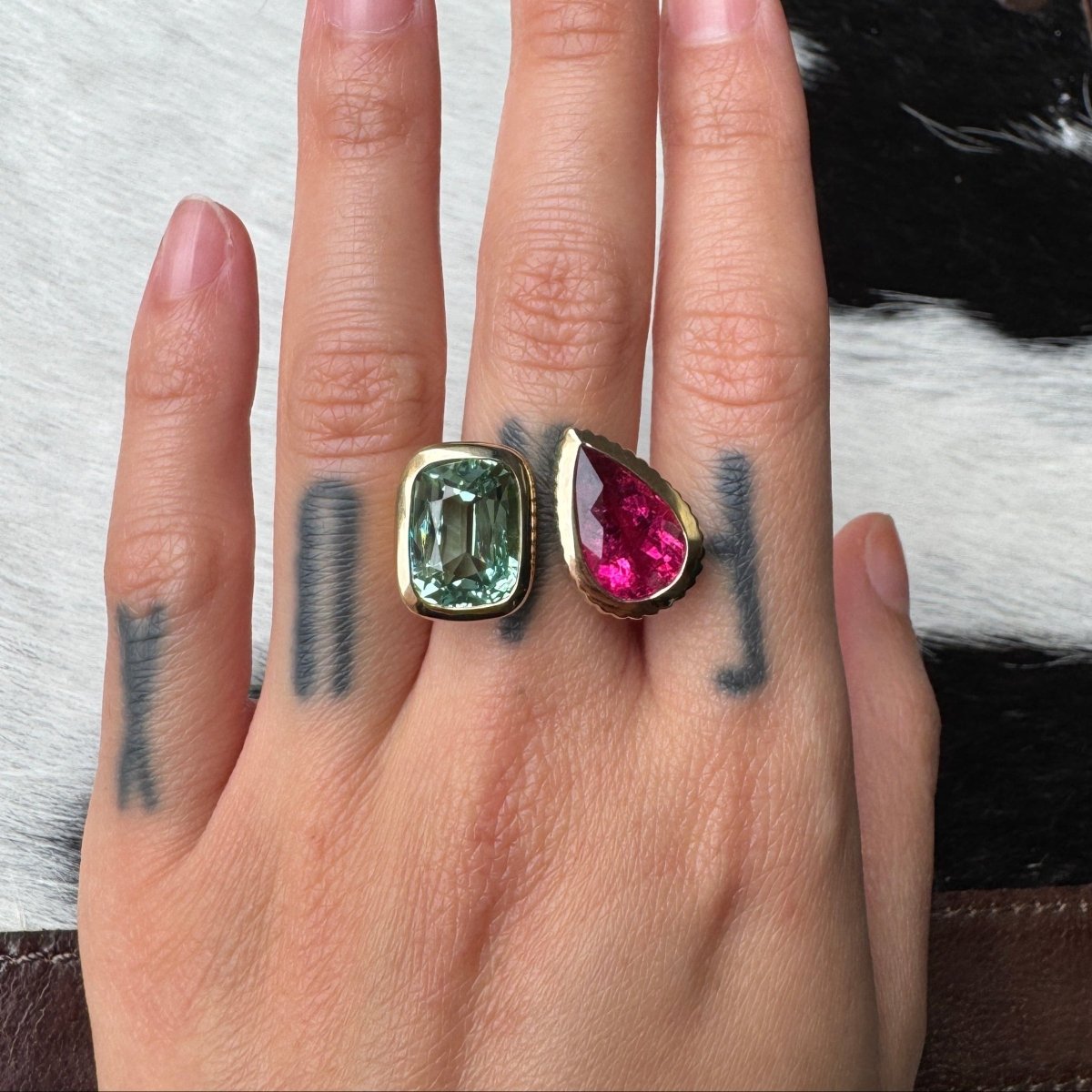 Hand wearing a gold ring with an elongated cushion-cut mint tourmaline and a pear-shaped pink red rubellite against a black and white cow print background