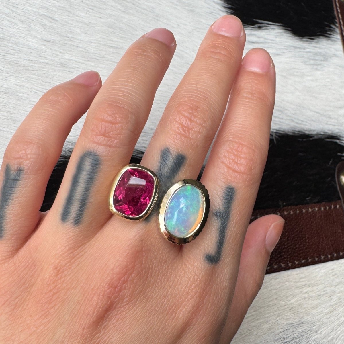 Hand wearing a gold ring with an elongated cushion-cut pink red rubellite and an oval cabochon rainbow opal against a black and white cow print background