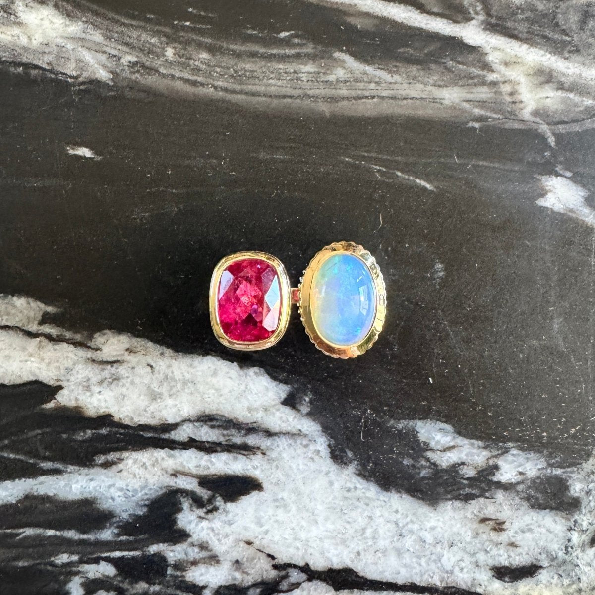 Gold ring with an elongated cushion-cut pink red rubellite and an oval cabochon rainbow opal against a black and white marble background 