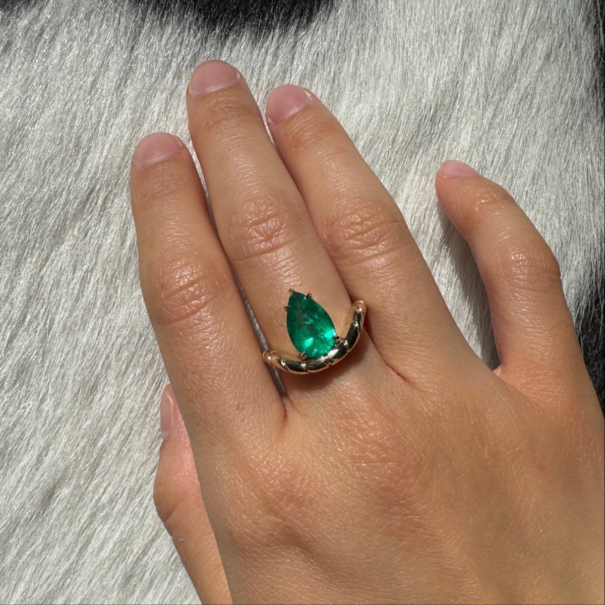 Hand wearing a gold ring with a curved band and a pear shaped emerald against a black and white cow print background