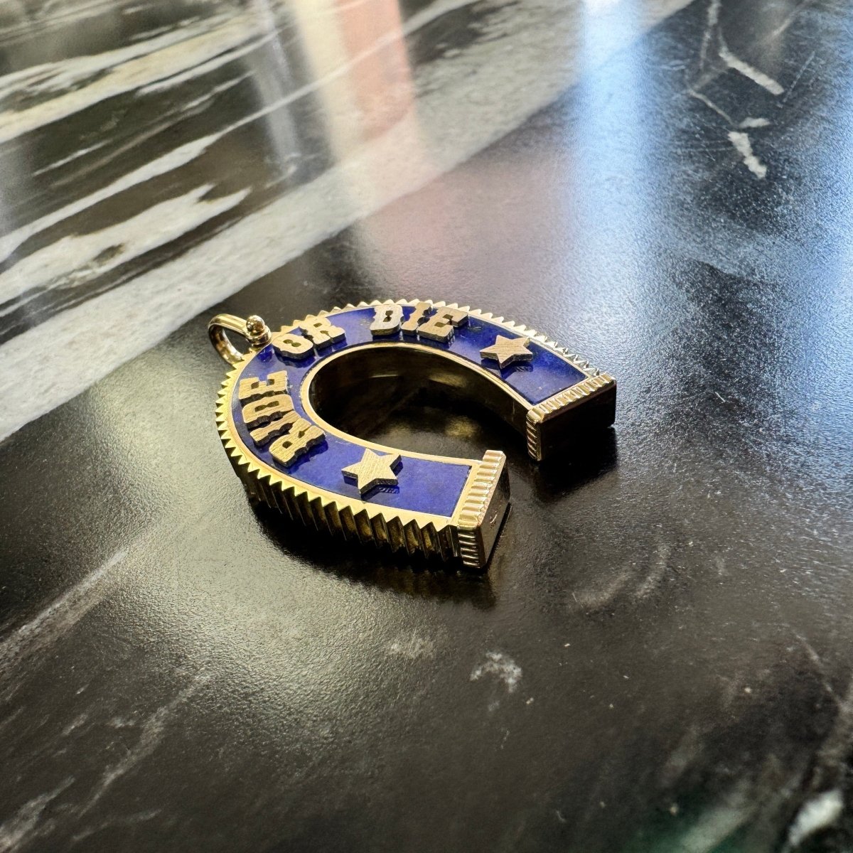 A gold horseshoe-shaped pendant with blue lapis and raised gold lettering spelling 'Ride or Die' with star details, resting on a dark marble surface.
