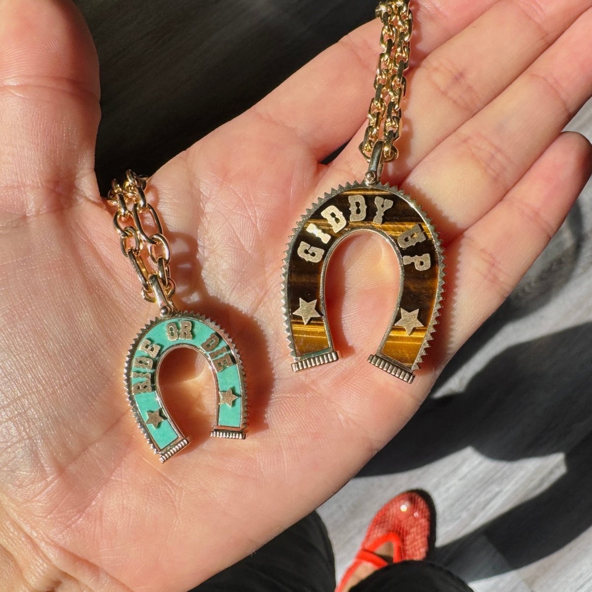 A hand holding two gold horseshoe-shaped necklaces or pendants with colorful inlay inserts, one turquoise and one gold with white lettering, over a gray wood floor.