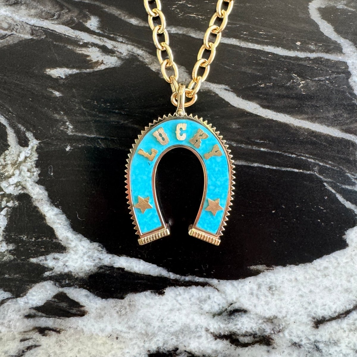 A gold chain necklace with a horseshoe-shaped pendant featuring turquoise blue enamel and the word 'LUCK' in gold lettering, displayed on a black and white marbled surface.