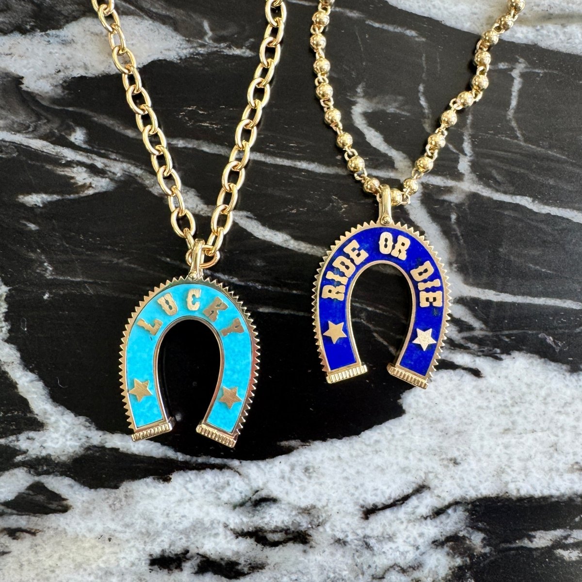 Two gold-tone horseshoe necklaces with colorful stone inlay pendants, one light blue with gold lettering and stars, and one dark blue with gold lettering and stars, displayed on a black and white marble surface.