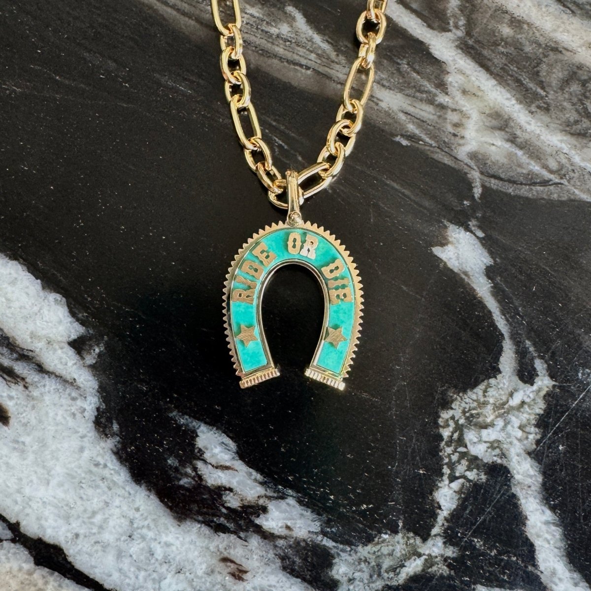 Gold chain necklace with a horseshoe-shaped pendant featuring turquoise and gold detailing, displayed on a black and white marble surface.