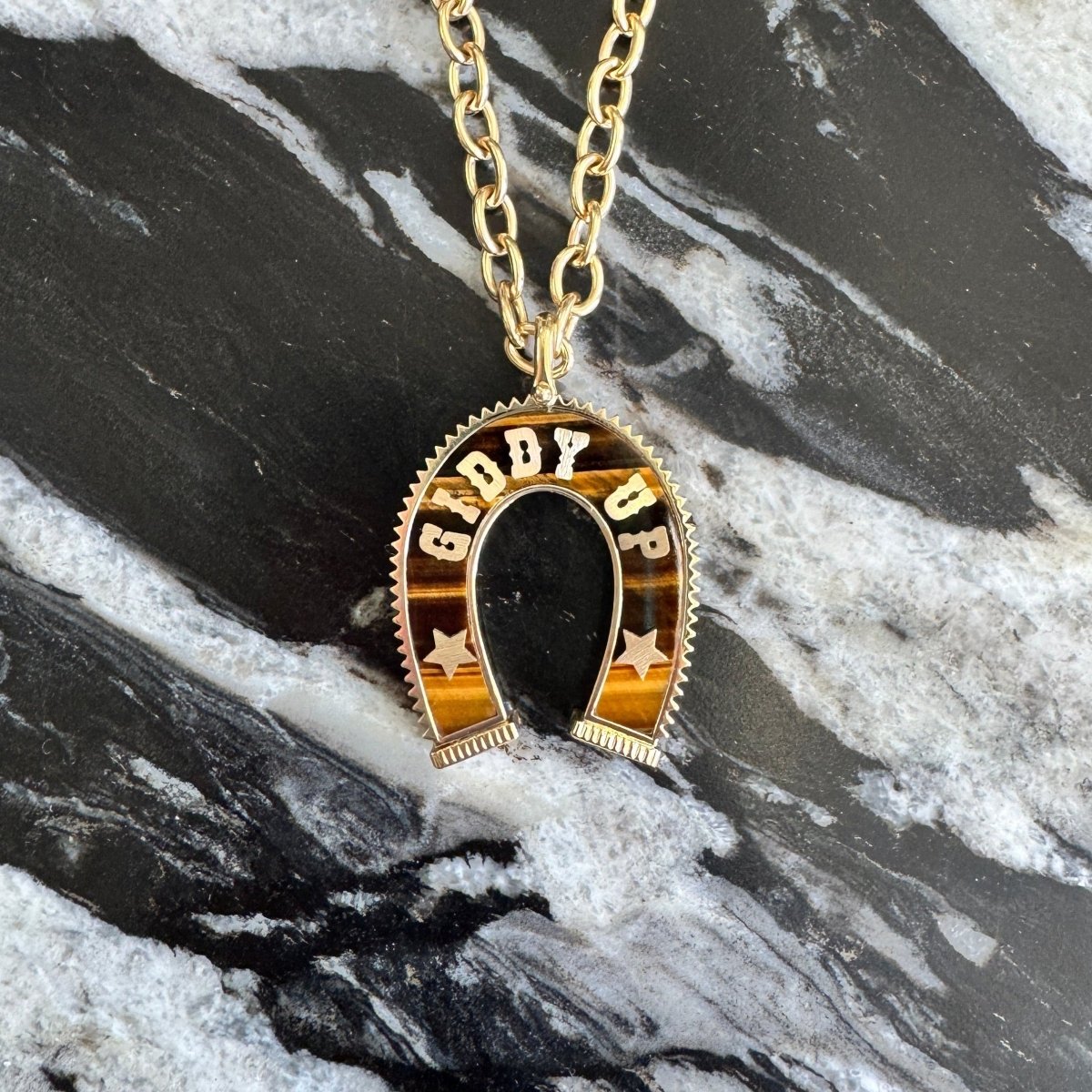 A gold horseshoe pendant necklace with 'GIDDY UP' and stars on a gold chain, displayed on a black and white marble surface.
