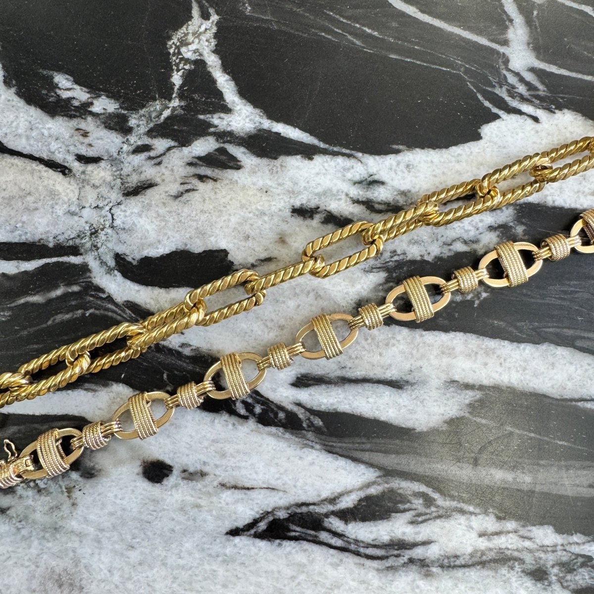 Multiple gold bracelets including a gold anchor link chain bracelet with rope detailing against a black and white marble background