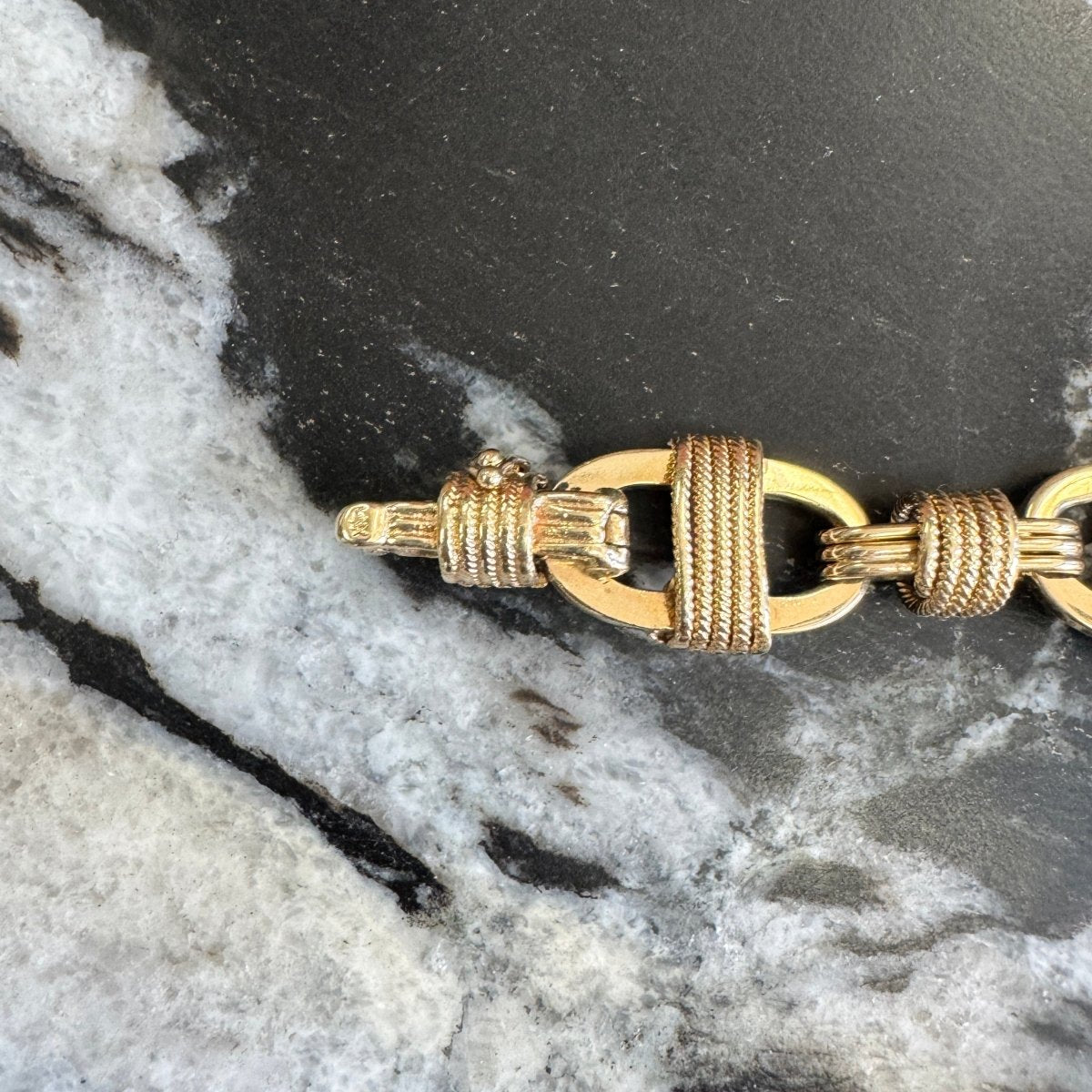 Close up of a gold clasp on an anchor link chain bracelet with rope detailing against a black and white marble background