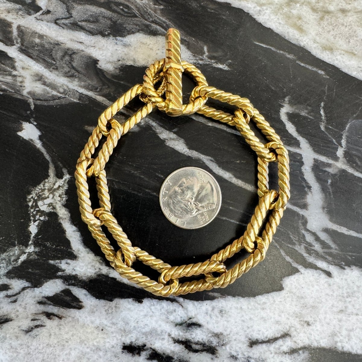 Gold oval link bracelet with rope texture near a quarter for size reference against a black and white marble background